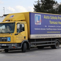 Auto Szkoła Premium - Żółta ciężarówka Auto Szkoła Premium z reklamą na naczepie, znak 'L', stożek drogowy, widok z boku, ulica miejska. Nauka jazdy kat. B, C, C+E, Taxi.