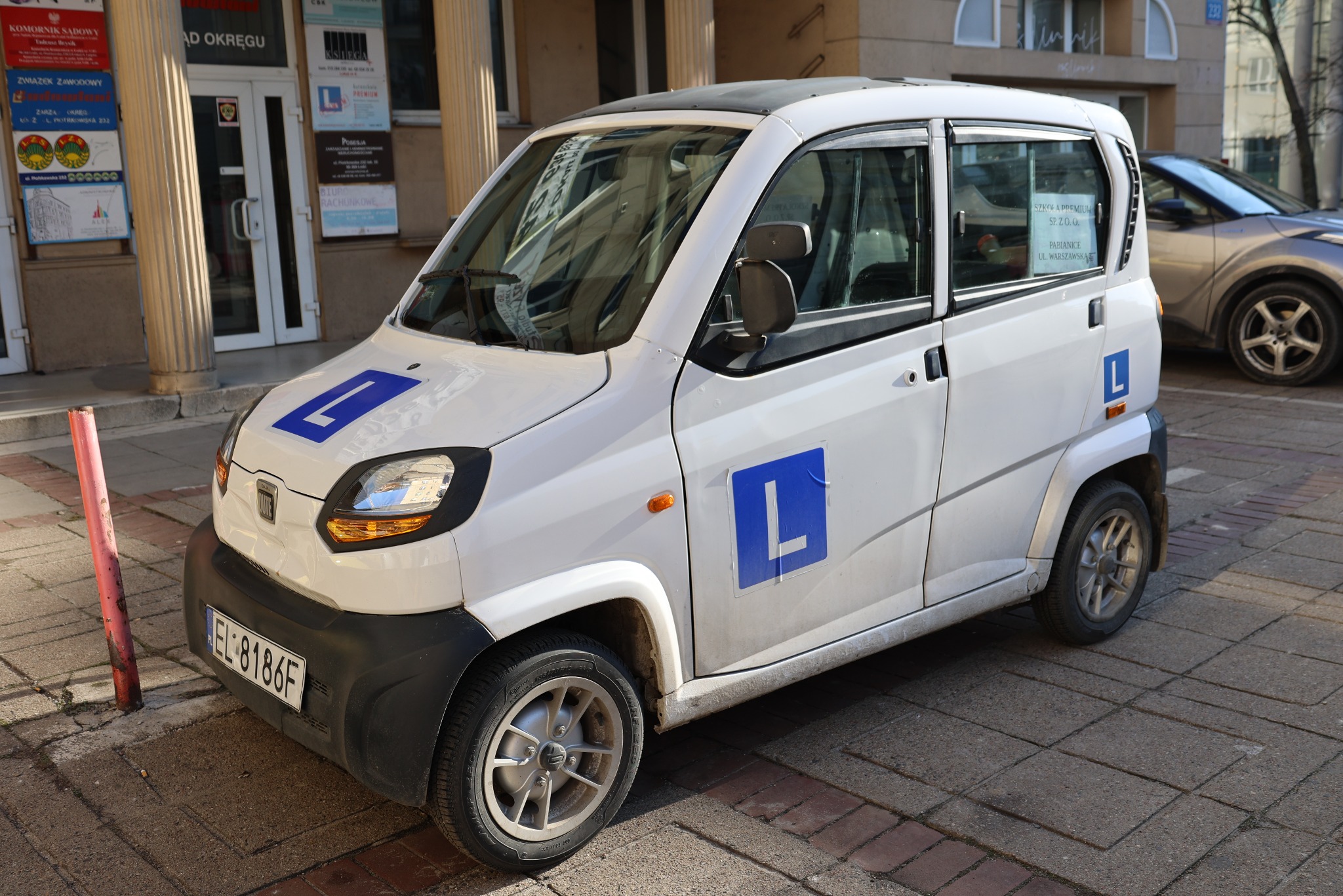 Biały samochód o nietypowym kształcie, oznaczony literą 'L', zaparkowany na chodniku przed budynkiem. Tablica 'Szkoła Premium' na szybie.