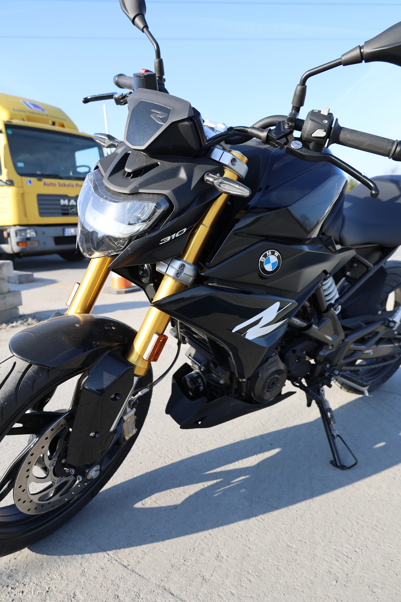 Czarny motocykl BMW G310R z żółtymi amortyzatorami przednimi, zaparkowany na placu manewrowym, w tle żółty samochód Auto Szkoły.