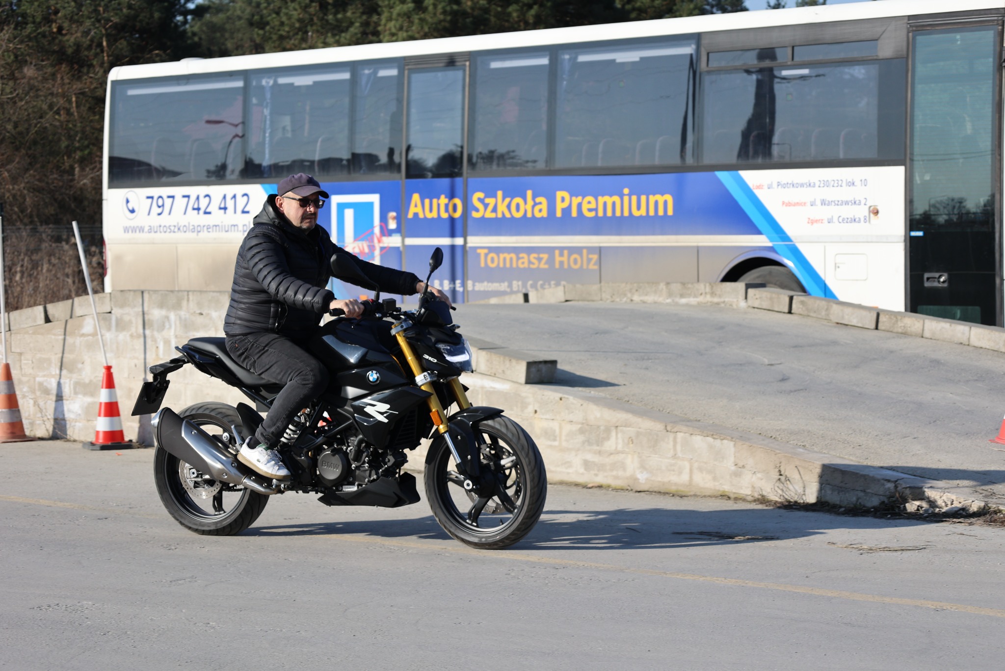 Kursant na motocyklu BMW 310, na tle autobusu Auto Szkoły Premium z danymi kontaktowymi. Jazda po placu manewrowym, widoczne pachołki.