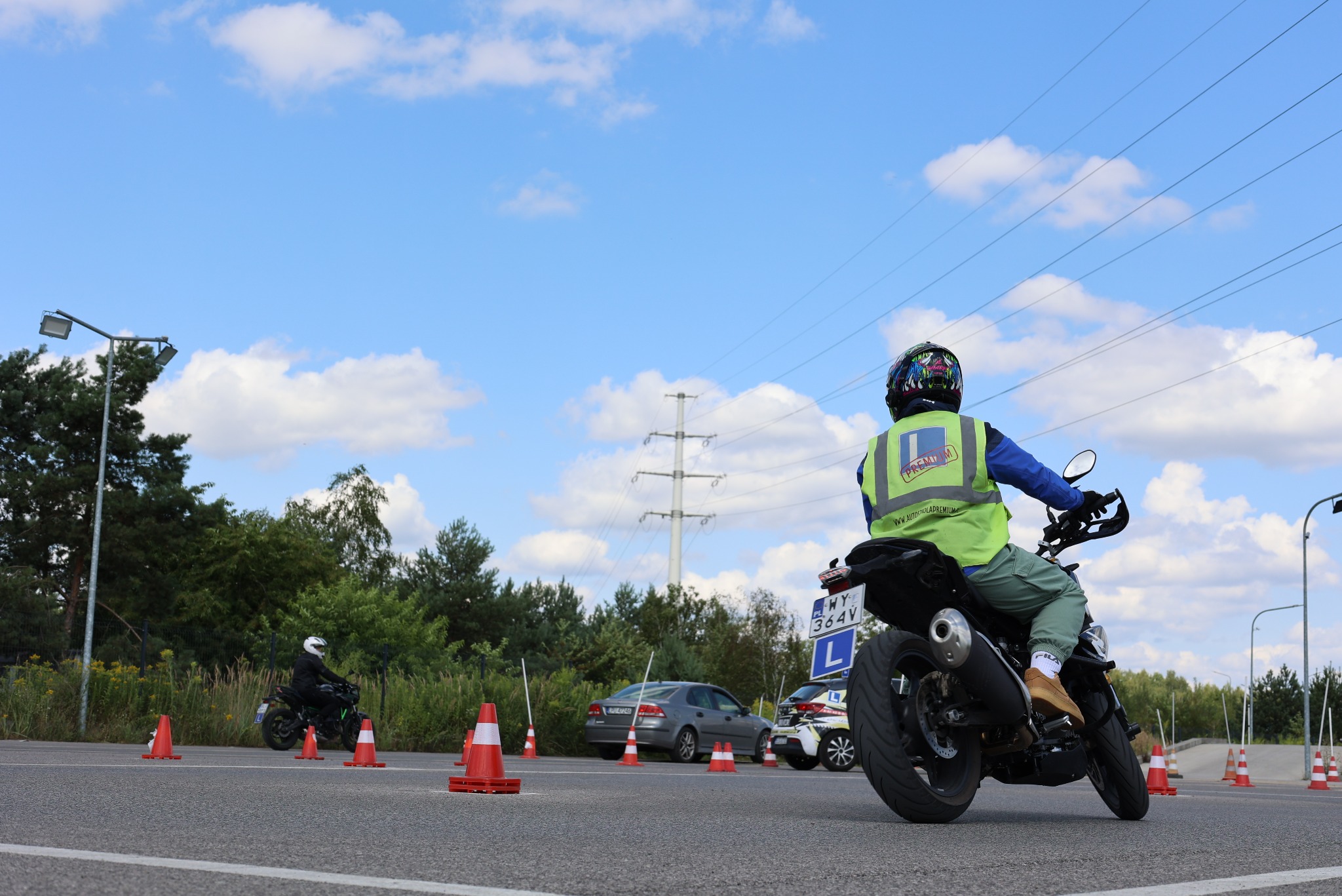 Kursant na motocyklu podczas szkolenia w szkole jazdy, widok z tyłu, pomarańczowe pachołki, niebieskie niebo z chmurami. Tablica z literą L na motocyklu.