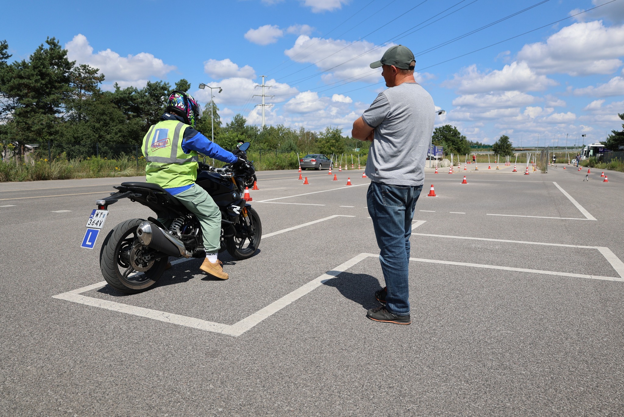 Instruktor obserwuje kursanta na motocyklu podczas egzaminu praktycznego na placu manewrowym z pachołkami i wyznaczonymi liniami.