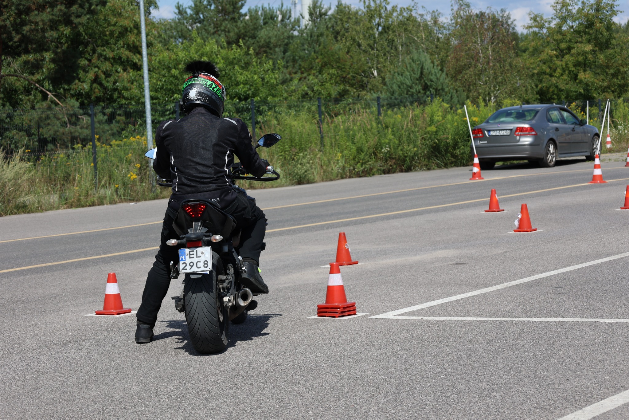 Motocyklista w kasku z pomponem, na placu manewrowym z pachołkami i liniami, podczas kursu prawa jazdy kat. A. Widok z tyłu, w tle auto i drzewa.