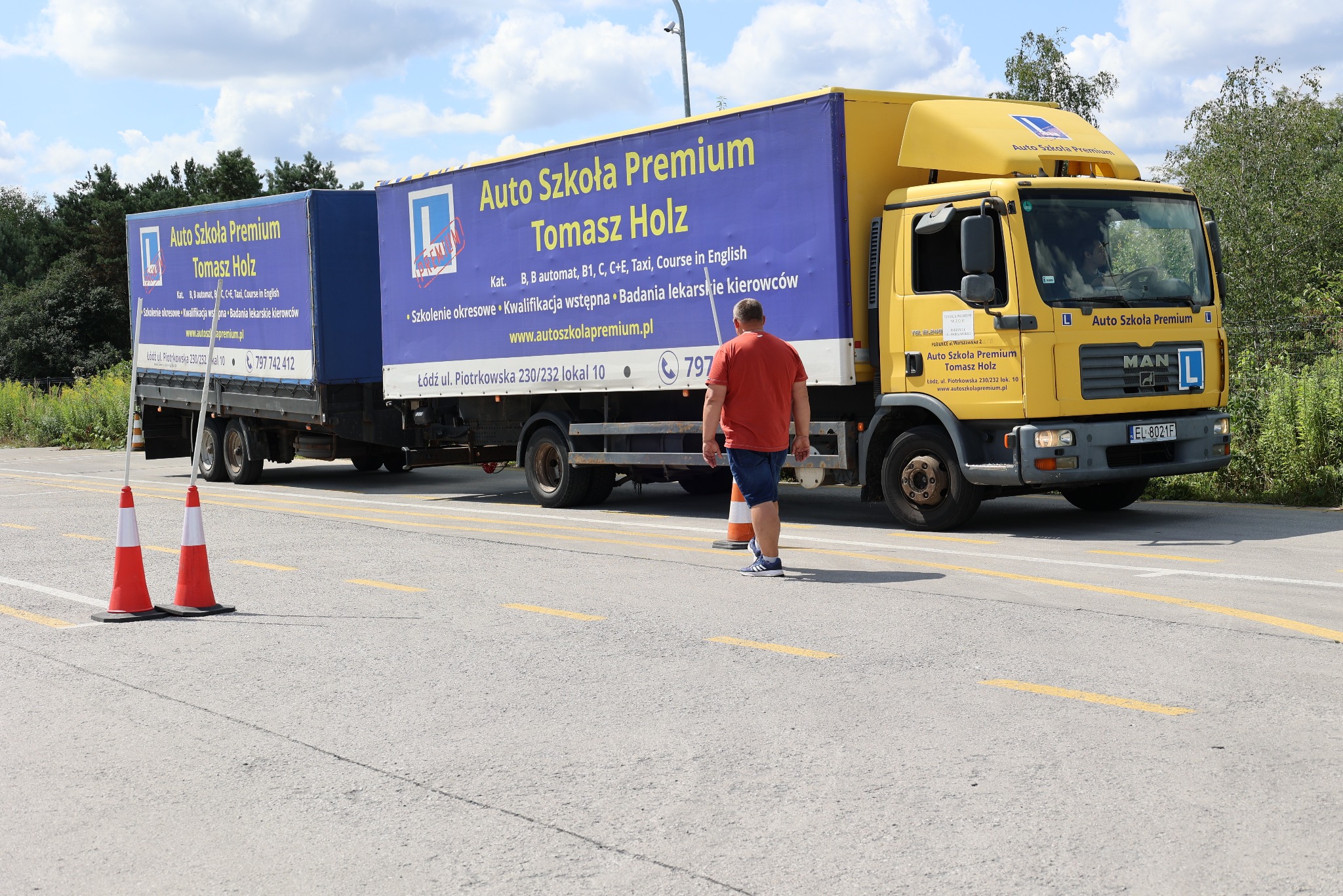 Żółta ciężarówka Auto Szkoły Premium z reklamą na naczepie, instruktor przy pachołku, asfaltowy plac manewrowy, drzewa w tle. Nauka jazdy kat. C+E.