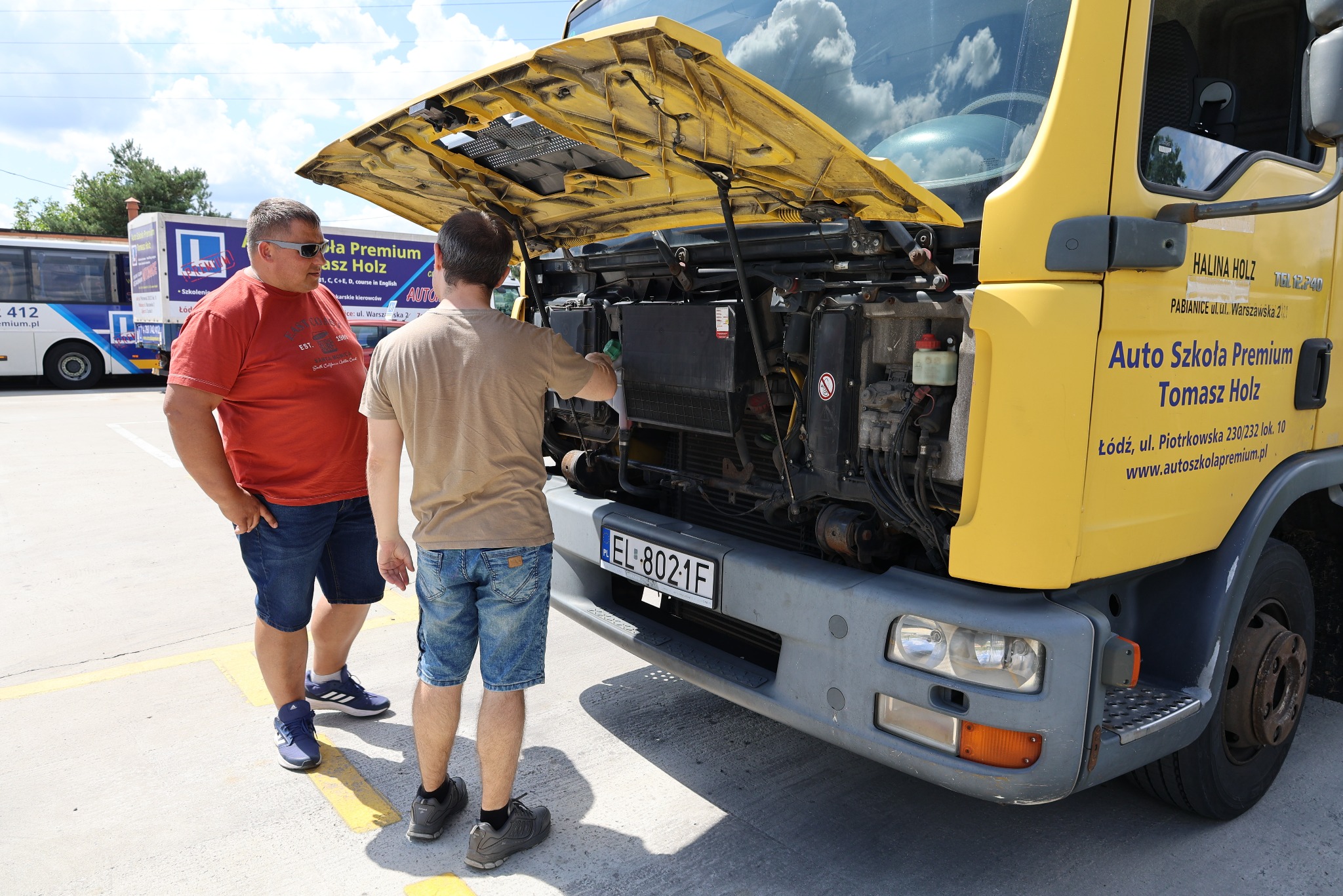 Instruktorzy auto szkoły Premium Tomasz Holz sprawdzają silnik żółtego samochodu ciężarowego, zaparkowanego na placu manewrowym.