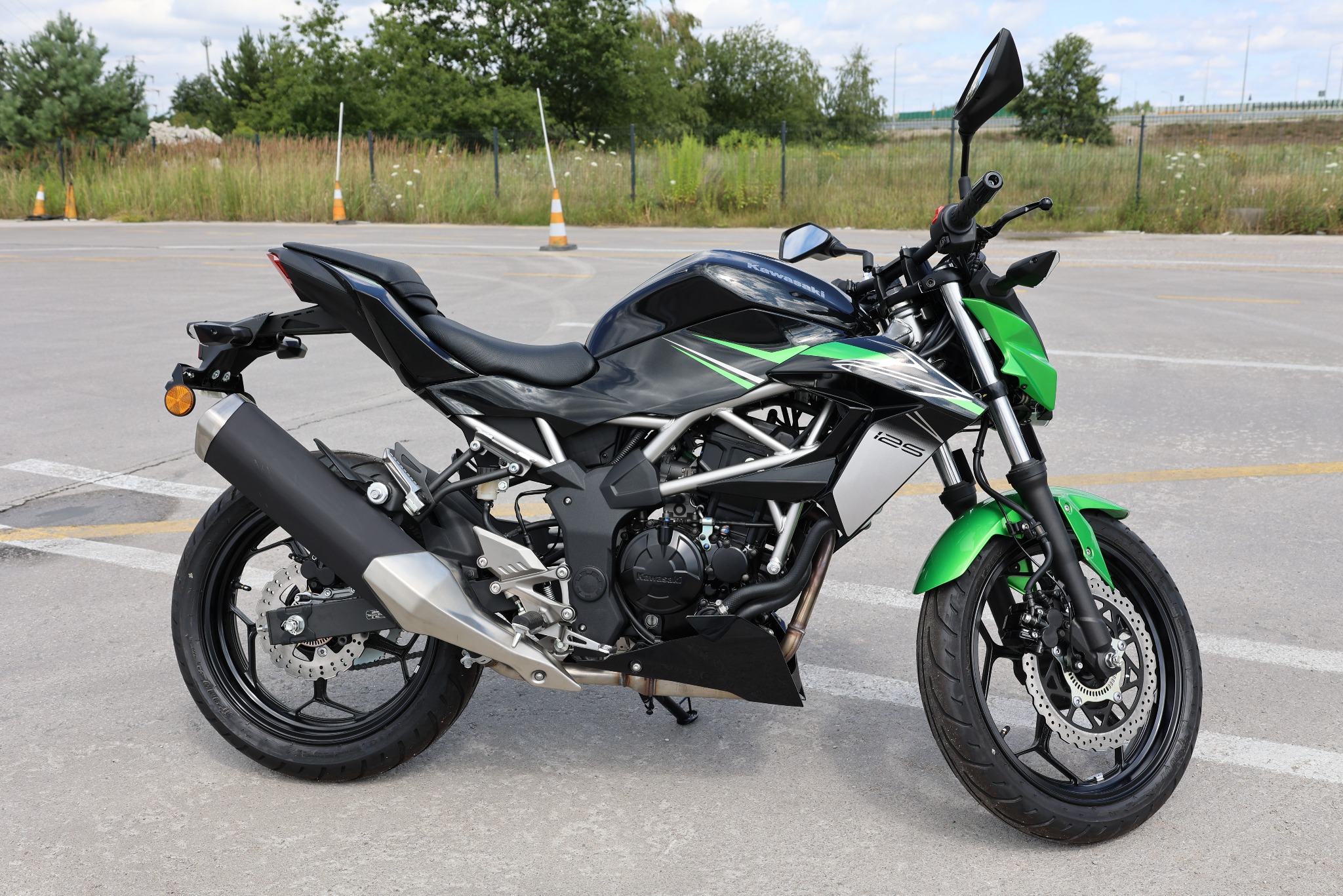 Czarno-zielony motocykl Kawasaki Ninja 125 na placu manewrowym z pomarańczowymi pachołkami w tle. Widok z boku, oświetlenie dzienne.
