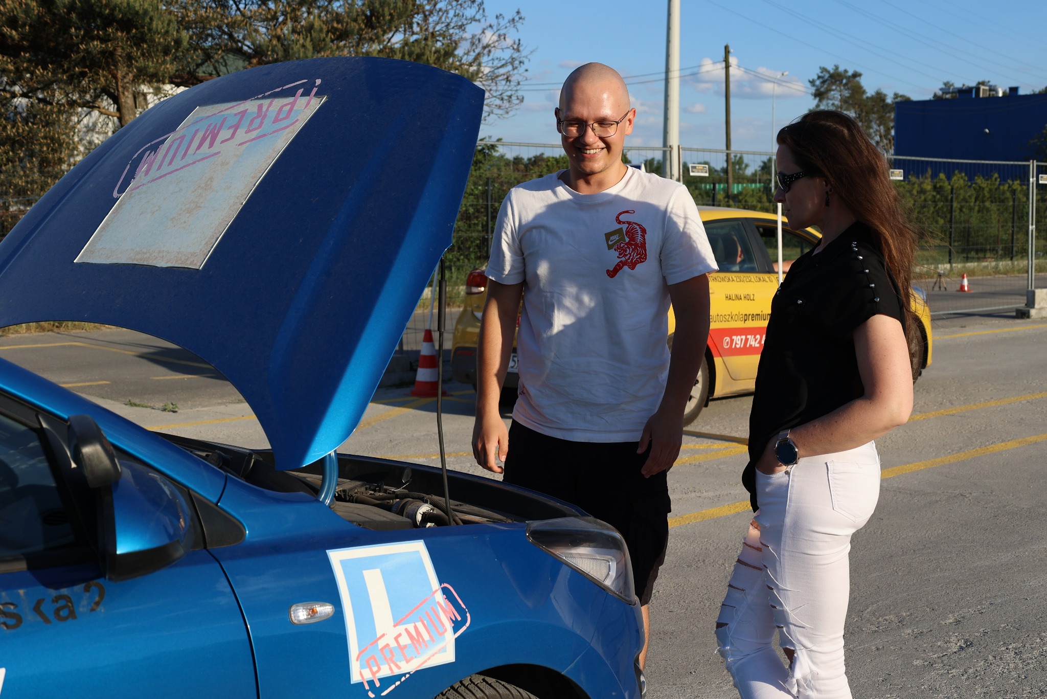 Instruktor z kursantką przy niebieskim aucie z otwartą maską i naklejką 'L' w Łodzi. Żółty samochód szkoły jazdy w tle.