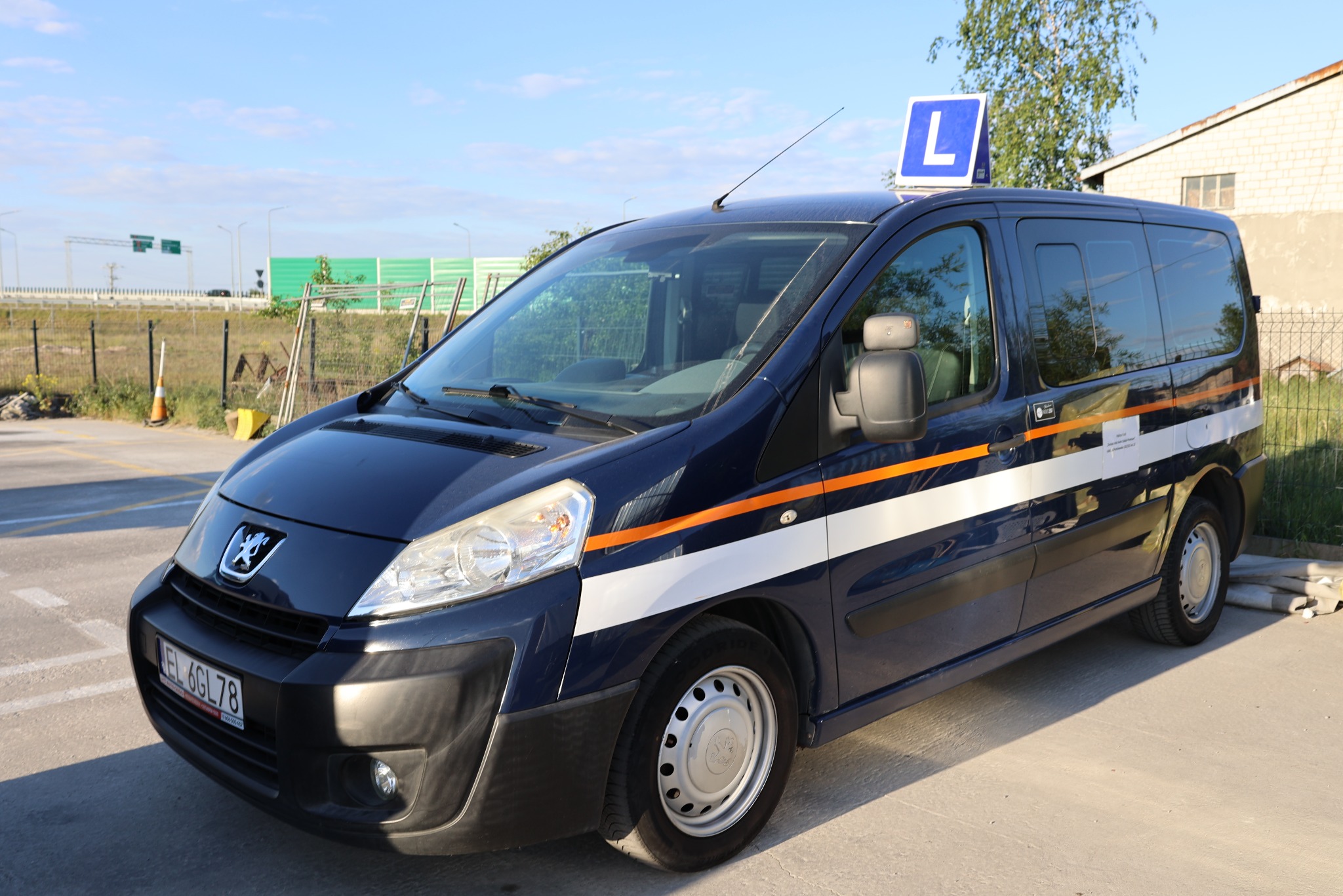 Niebieski Peugeot z pomarańczowymi pasami i literą 'L' na dachu, zaparkowany na placu manewrowym szkoły jazdy, w tle widoczna infrastruktura drogowa.