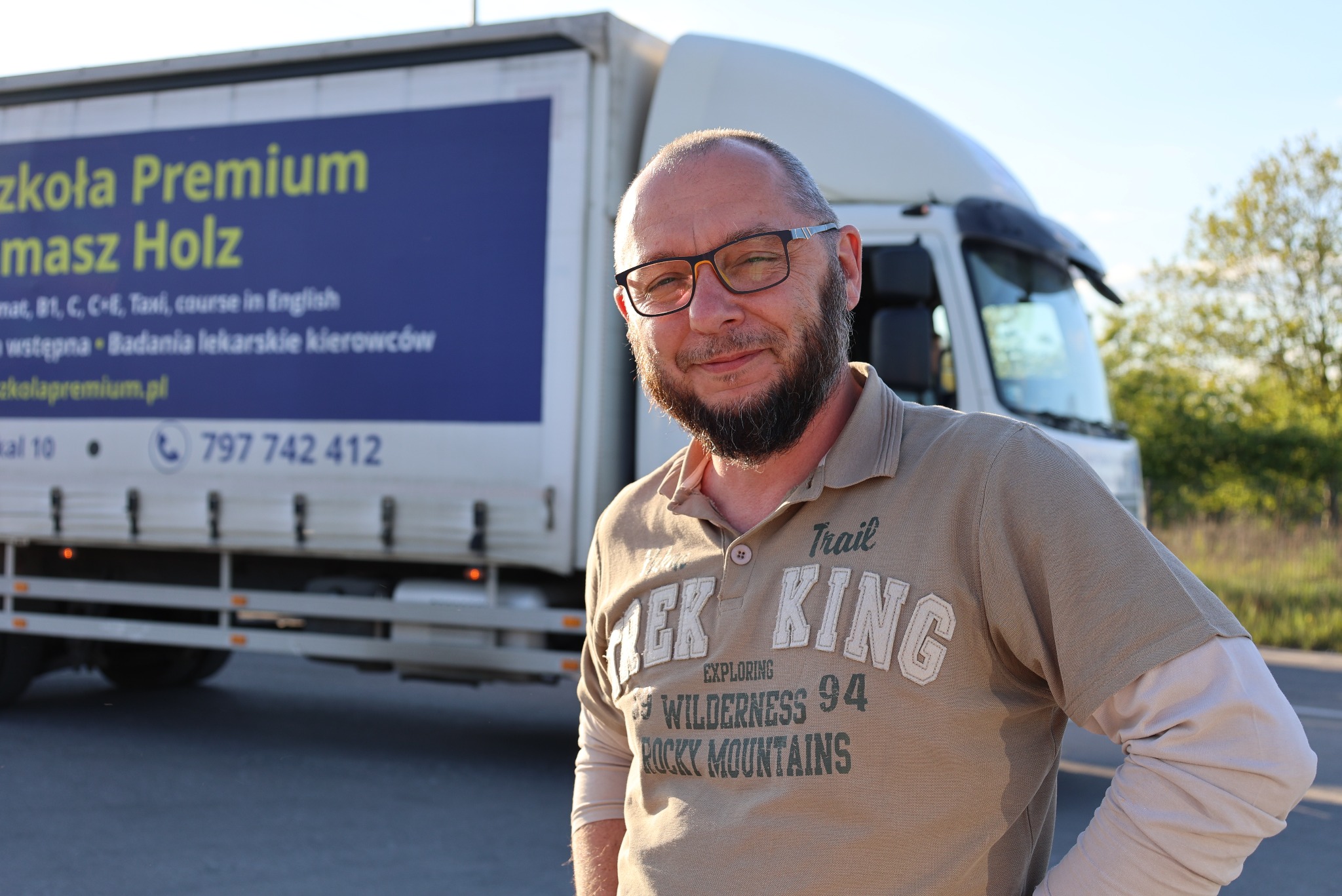 Portret mężczyzny w okularach na tle ciężarówki Szkoły Premium Tomasz Holz, oferującej kursy kat. B, C, C+E, Taxi i badania lekarskie. Kontakt: 797 742 412.