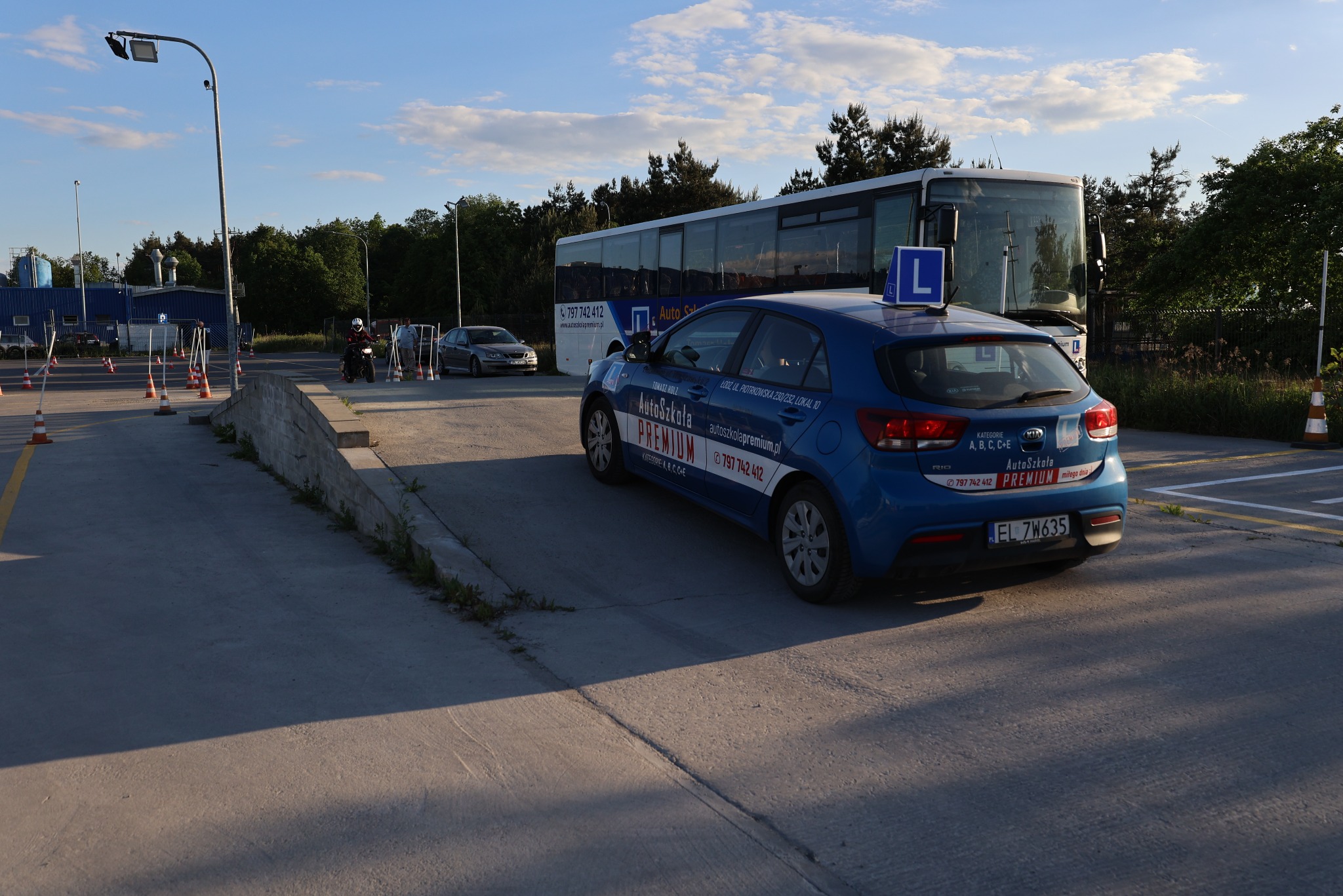 Niebieski samochód z literą 'L' na dachu, autobus i plac manewrowy szkoły jazdy AutoSzkoła Premium. Widoczne logo i numer telefonu na pojazdach.