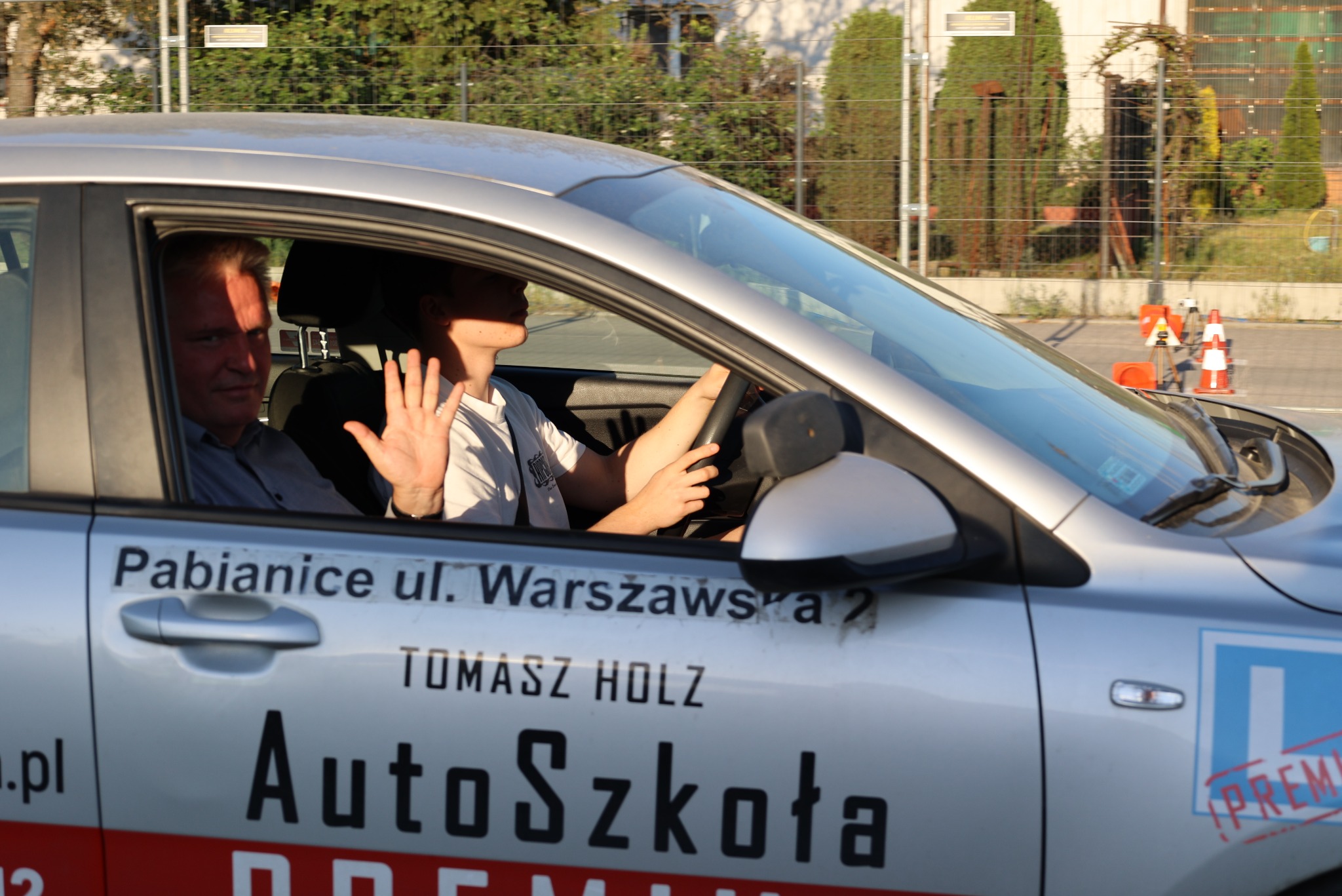 Auto szkoły z instruktorem i kursantem w trakcie jazdy. Na drzwiach napis 'Auto Szkoła' i informacja o lokalizacji: Pabianice, ul. Warszawska.