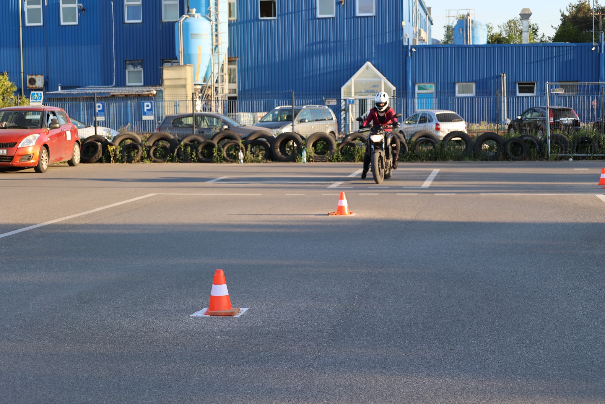 Kobieta na motocyklu pokonuje tor przeszkód z pachołkami na placu manewrowym szkoły jazdy. W tle budynek i opony.