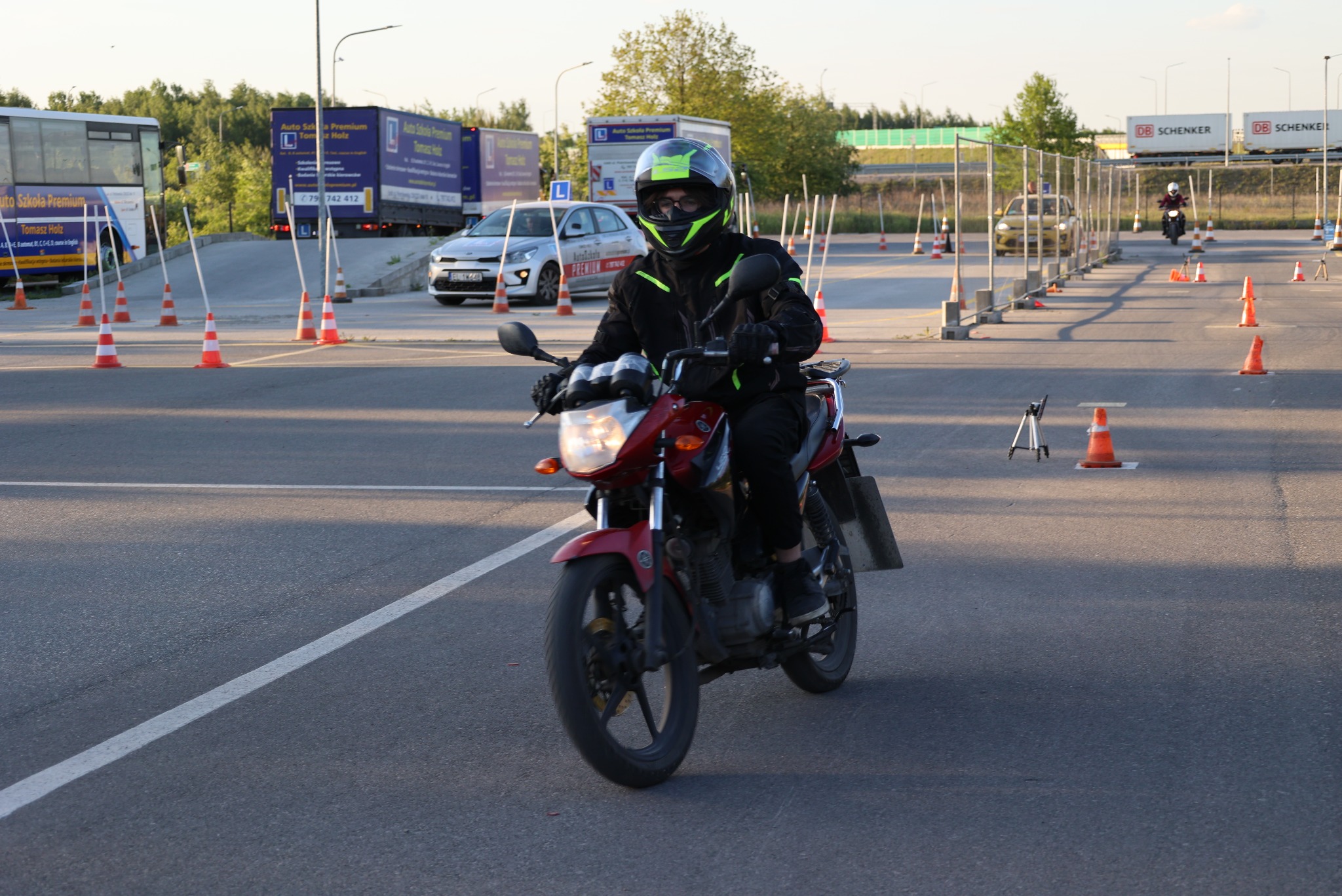 Kursant w kasku i kurtce na motocyklu, ćwiczący manewry na placu Szkoły Jazdy Premium z pomarańczowymi pachołkami i pojazdami w tle.