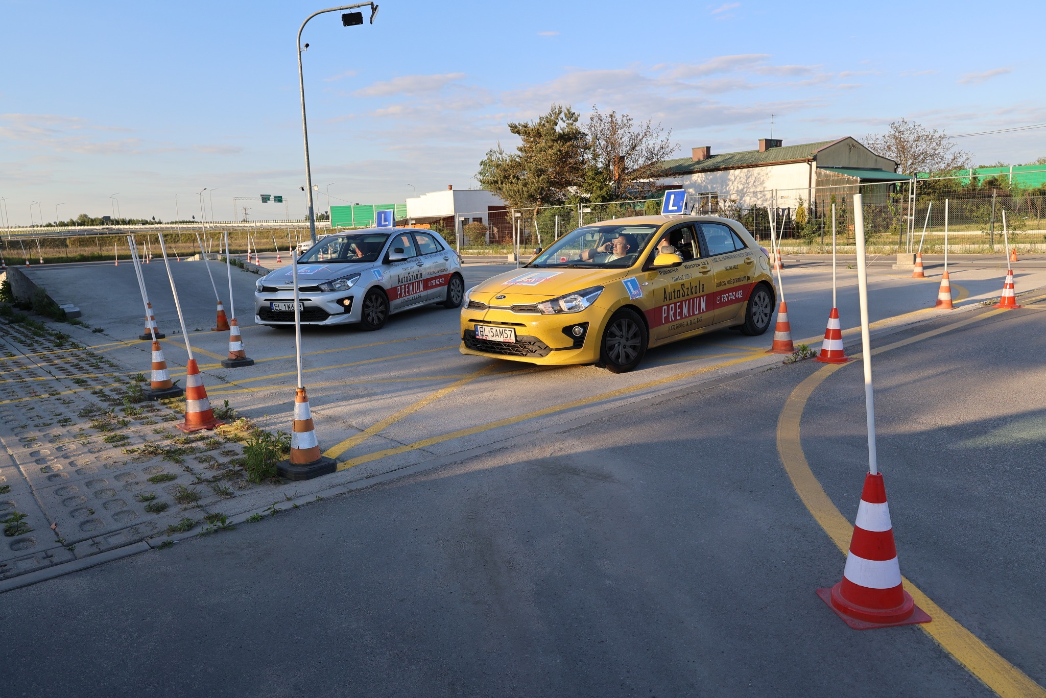 Dwa samochody szkoły jazdy z kursantami na placu manewrowym, otoczone pachołkami. Żółty i srebrny pojazd z oznaczeniem 'L' na dachu. Słoneczny dzień.