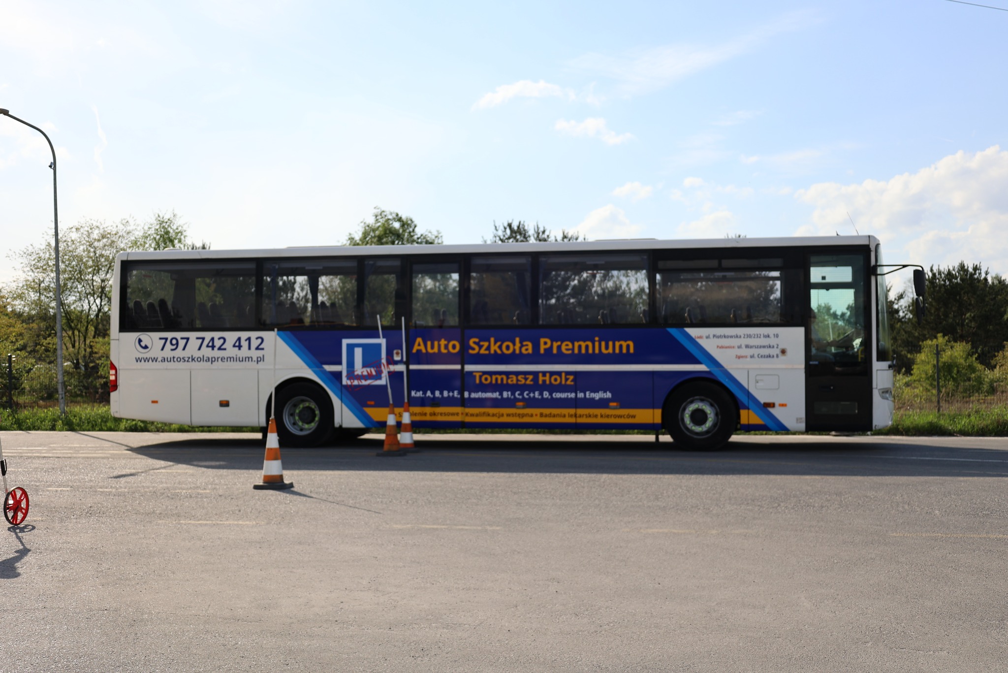Autobus Auto Szkoła Premium z numerem telefonu i adresem strony internetowej, pomarańczowe pachołki na asfalcie, w tle zieleń. Kursy kat. A, B, C, D, B+E.