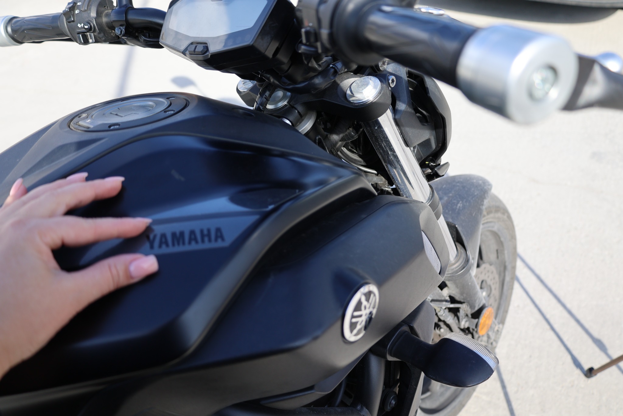 Czarny motocykl Yamaha z bliska, dłoń kobiety dotyka baku paliwa. Widoczne detale: kierownica, przednie koło i logo marki. Jasne, naturalne oświetlenie.