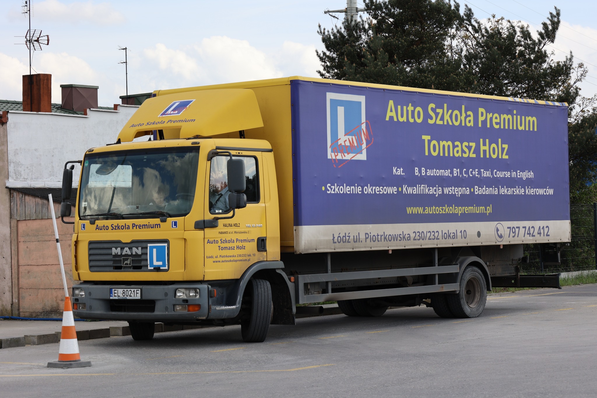 Żółta ciężarówka Auto Szkoła Premium z reklamą na naczepie, znak 'L', stożek drogowy, widok z boku, ulica miejska. Nauka jazdy kat. B, C, C+E, Taxi.