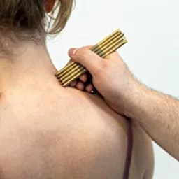 Masażer z drewna na karku pacjentki podczas zabiegu. Widoczna ręka terapeuty, jasne tło. Ujęcie z bliska, skupione na obszarze zabiegu.