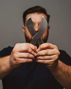 Mężczyzna trzyma skrzyżowane narzędzia Gua Sha przed twarzą. Szare tło, zbliżenie na dłonie i narzędzia, ciemne ubranie. Nietypowe ujęcie, brak widocznych rysów twarzy.
