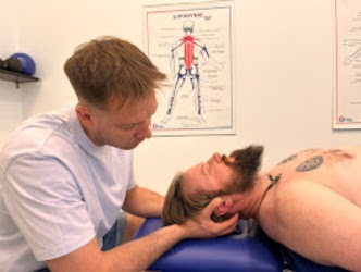 Terapeuta manualny delikatnie manipuluje szyją pacjenta leżącego na niebieskim stole; w tle plakat anatomiczny i sprzęt do ćwiczeń.