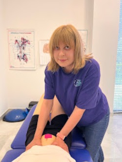 Fizjoterapeutka w fioletowej koszulce wykonuje terapię manualną bioder pacjentki leżącej na niebieskim stole do masażu, widoczne plakaty anatomiczne w tle.