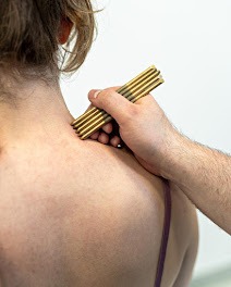 Masażer z drewna na karku pacjentki podczas zabiegu. Widoczna ręka terapeuty, jasne tło. Ujęcie z bliska, skupione na obszarze zabiegu.