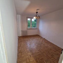 Dom Pro Bud - Pusty pokój z parkietem w jodełkę, świeżo pomalowane ściany na biało, okno z widokiem na zieleń. Widoczny grzejnik i lampa sufitowa.