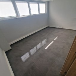 Dom Pro Bud - Wykończone wnętrze: szare płytki podłogowe z połyskiem, białe ściany i listwy przypodłogowe, okno z roletami. Widoczne gniazdka elektryczne. Nowoczesny minimalizm.