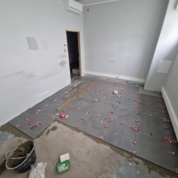 Dom Pro Bud - Szare płytki podłogowe w trakcie układania, z czerwonymi krzyżykami dystansowymi, poziomica, wiadro z zaprawą, w surowym wnętrzu.