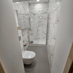 Nowoczesna łazienka po remoncie: białe płytki imitujące marmur, szara podłoga, kabina prysznicowa ze szklanymi drzwiami i podwieszana toaleta. Widok zza futryny.