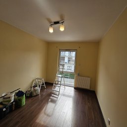 Dom Pro Bud - Pokój po malowaniu na żółto, widoczne drabiny, pędzle i farby. Podłoga z paneli, okno z widokiem na budynek. Przygotowanie do dalszych prac.