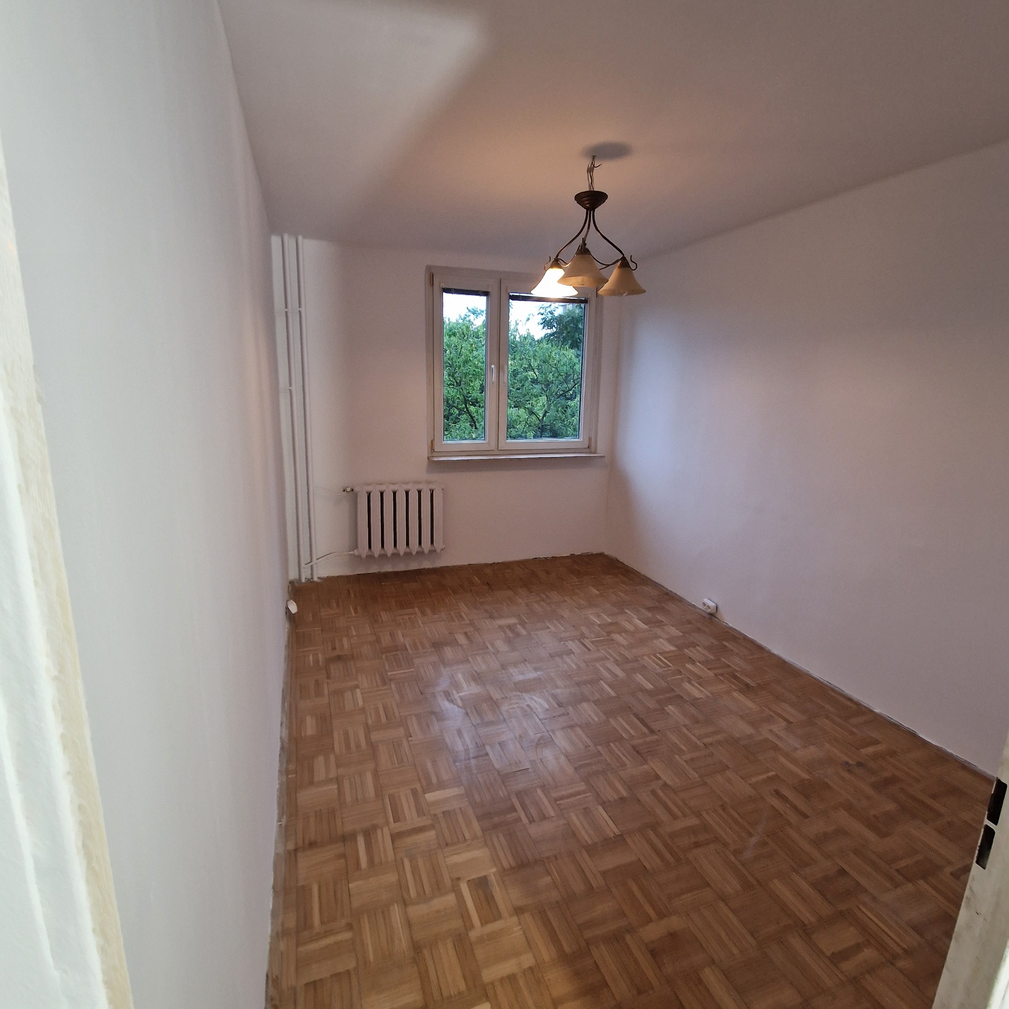 Pusty pokój z parkietem w jodełkę, świeżo pomalowane ściany na biało, okno z widokiem na zieleń. Widoczny grzejnik i lampa sufitowa.