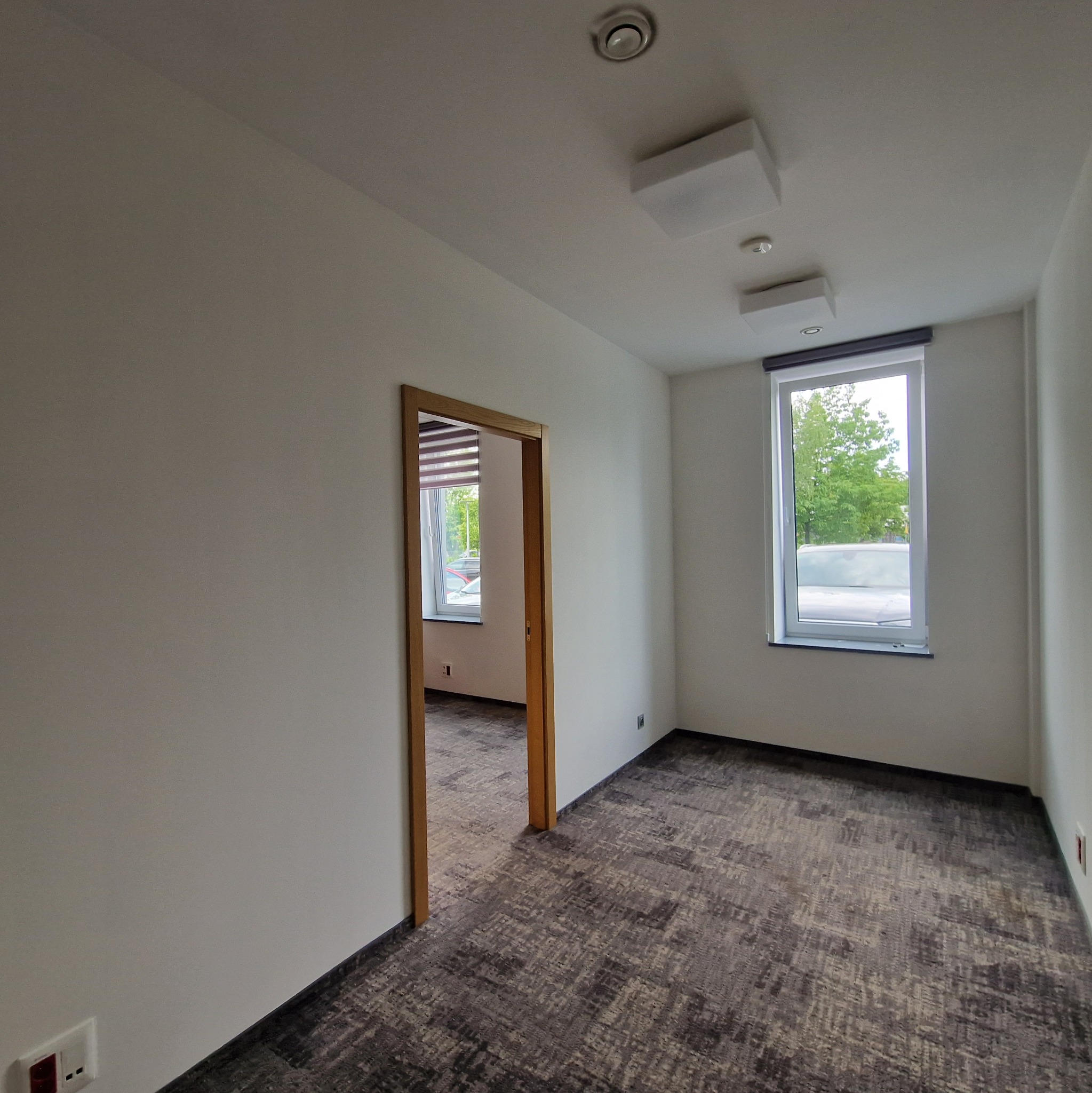 Jasne, puste pomieszczenie biurowe z oknem i widokiem na zieleń. Wnętrze po remoncie, z nową podłogą i świeżo pomalowanymi ścianami. Widok na sąsiednie pomieszczenie.