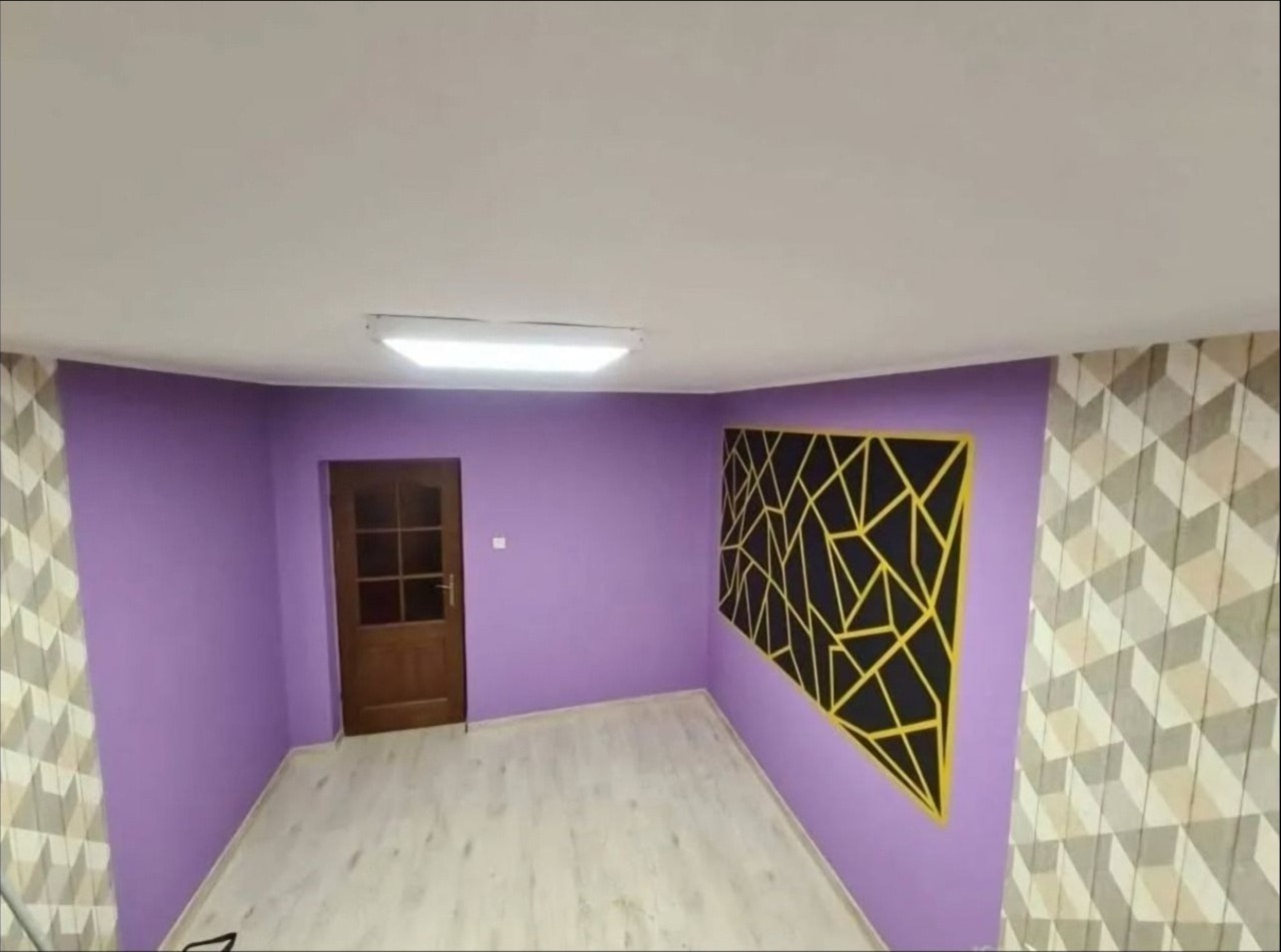 Wnętrze pokoju po malowaniu ścian na fioletowo, z drewnianymi drzwiami i dekoracją geometryczną na ścianie. Podłoga jasna, panelowa. Jedna ze ścian pokryta tapetą.