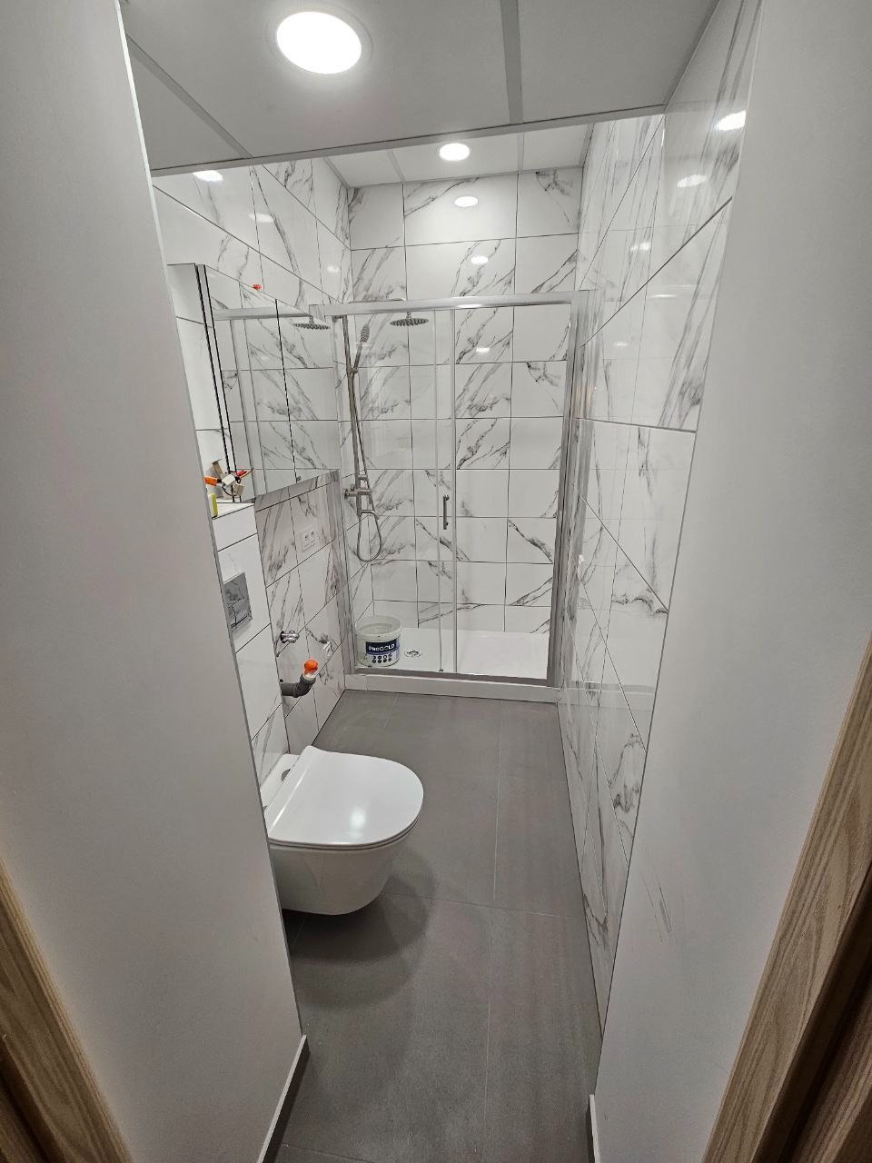 Nowoczesna łazienka po remoncie: białe płytki imitujące marmur, szara podłoga, kabina prysznicowa ze szklanymi drzwiami i podwieszana toaleta. Widok zza futryny.