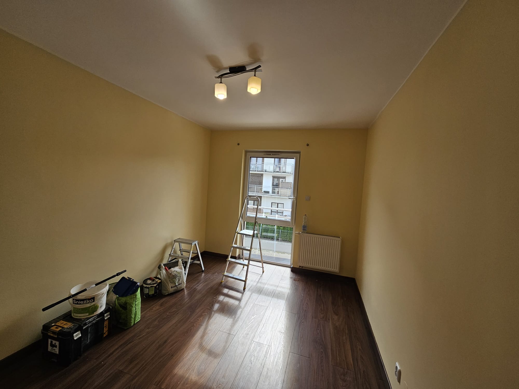 Pokój po malowaniu na żółto, widoczne drabiny, pędzle i farby. Podłoga z paneli, okno z widokiem na budynek. Przygotowanie do dalszych prac.