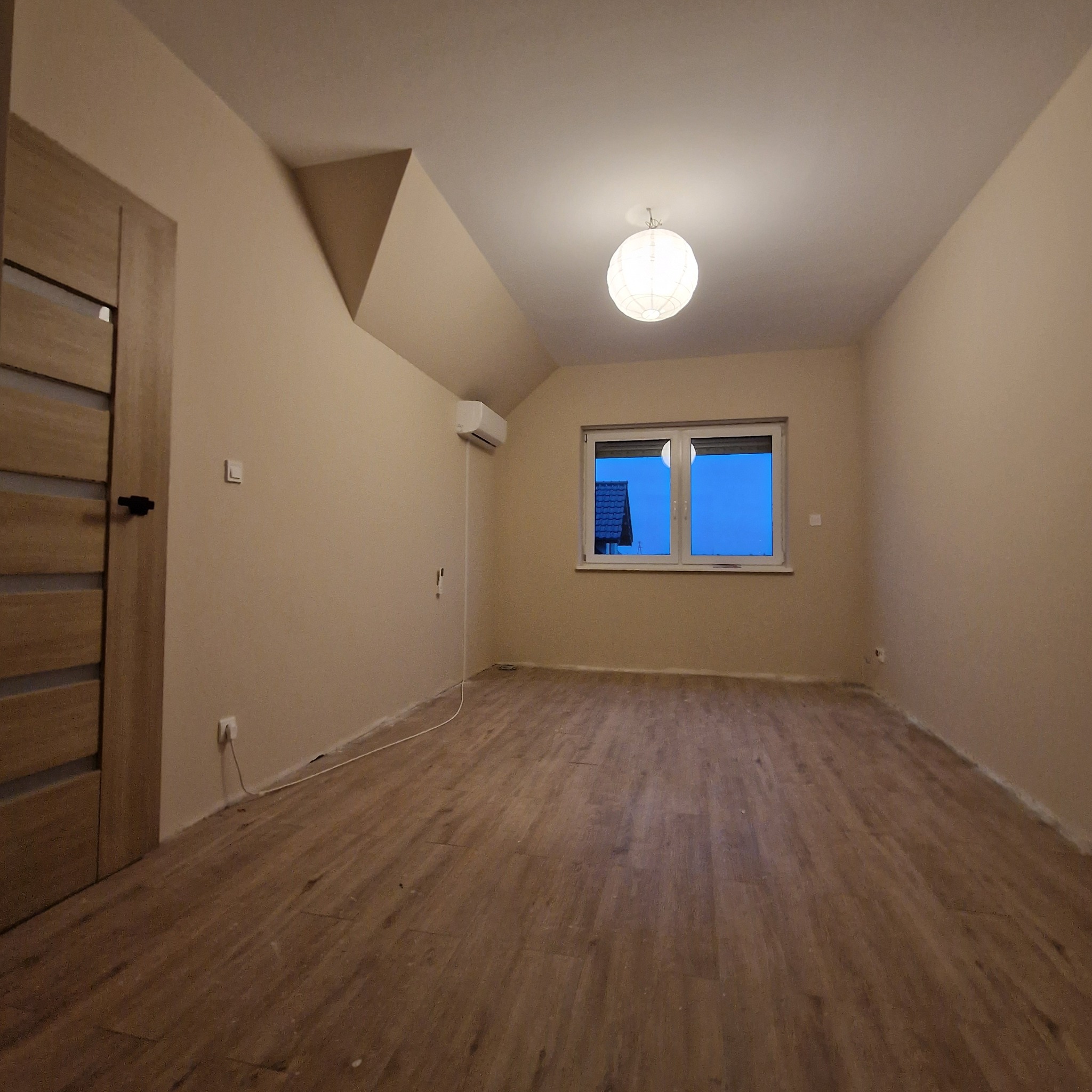 Jasne, puste wnętrze poddasza z oknem, drewnianą podłogą i świeżo pomalowanymi ścianami w odcieniach beżu. Widoczny klimatyzator i lampa sufitowa.