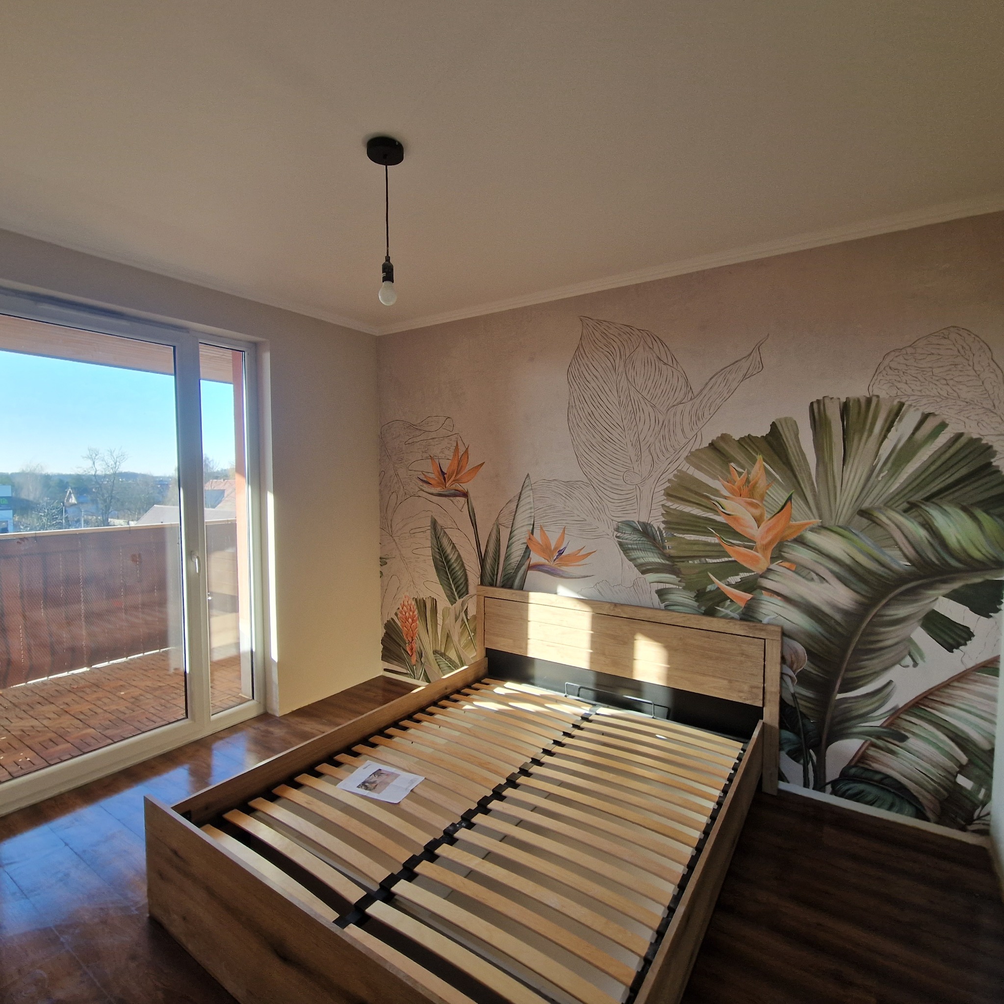 Sypialnia po remoncie: łóżko z drewnianą ramą na tle ściany z motywem tropikalnych roślin. Widok na balkon i jasne, naturalne światło.