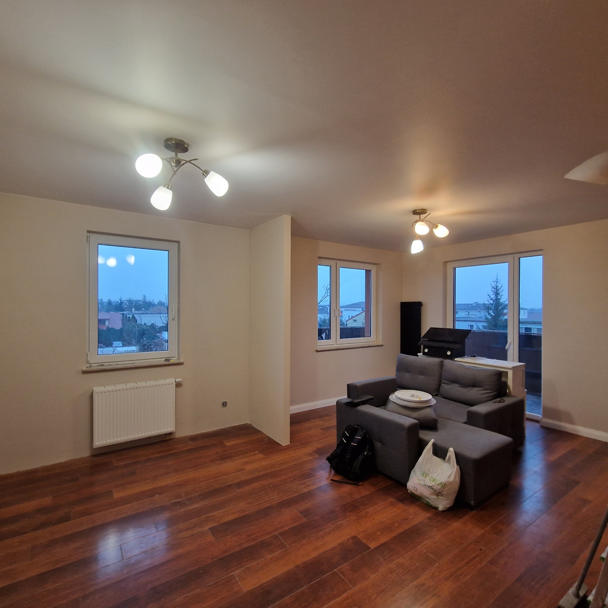 Jasne wnętrze po remoncie: drewniana podłoga, szara sofa, okna z widokiem na zewnątrz. Nowoczesne lampy sufitowe. Torba na podłodze dodaje realizmu.