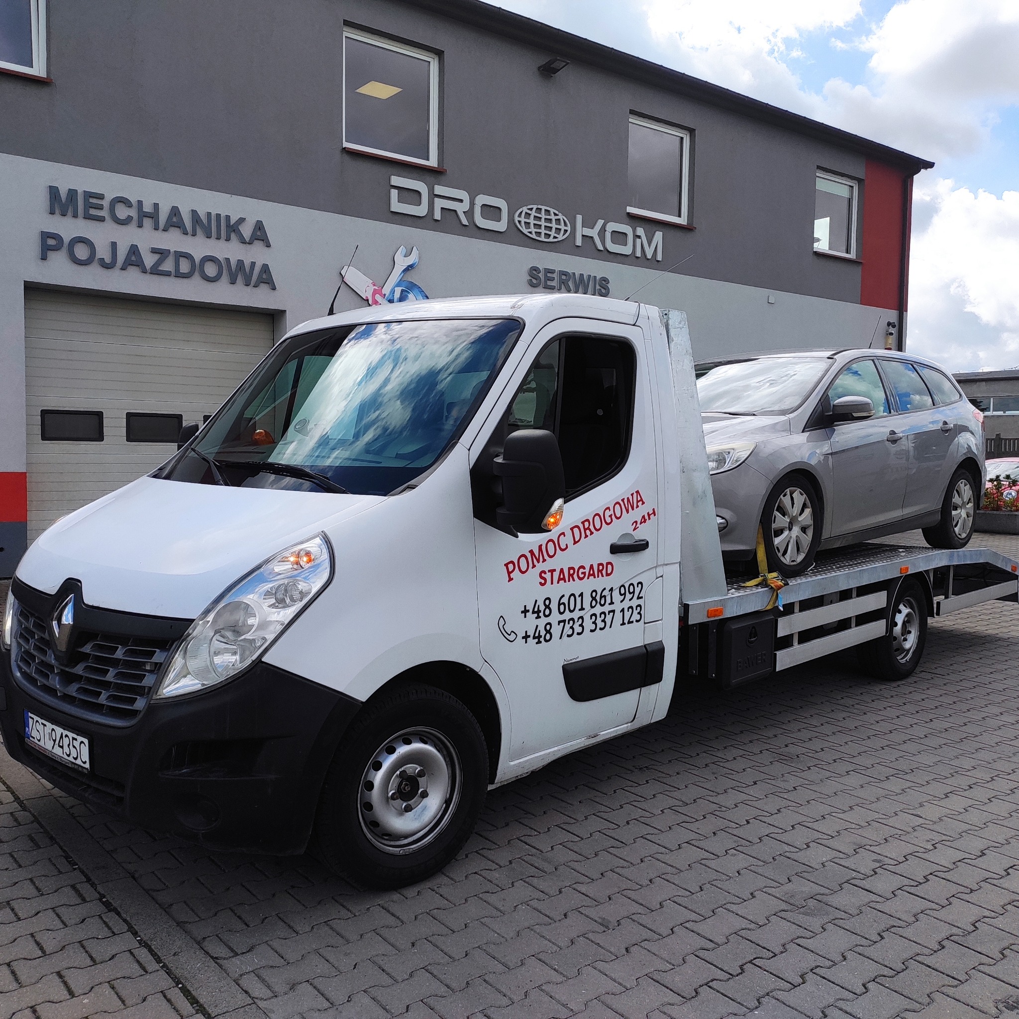 Laweta z szarym samochodem na platformie przed budynkiem warsztatu samochodowego Drok-om Serwis. Pomoc drogowa Stargard 24h.