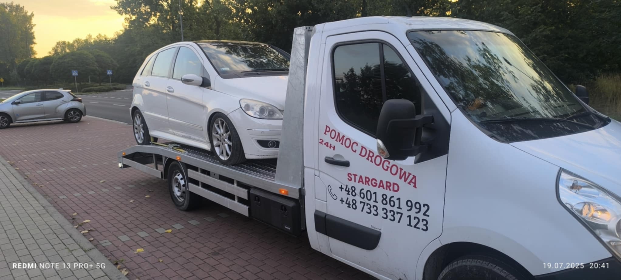 Laweta z białym Mercedesem na platformie, na boku napis 'Pomoc Drogowa 24H Stargard' i numery telefonów. W tle zaparkowany szary samochód.