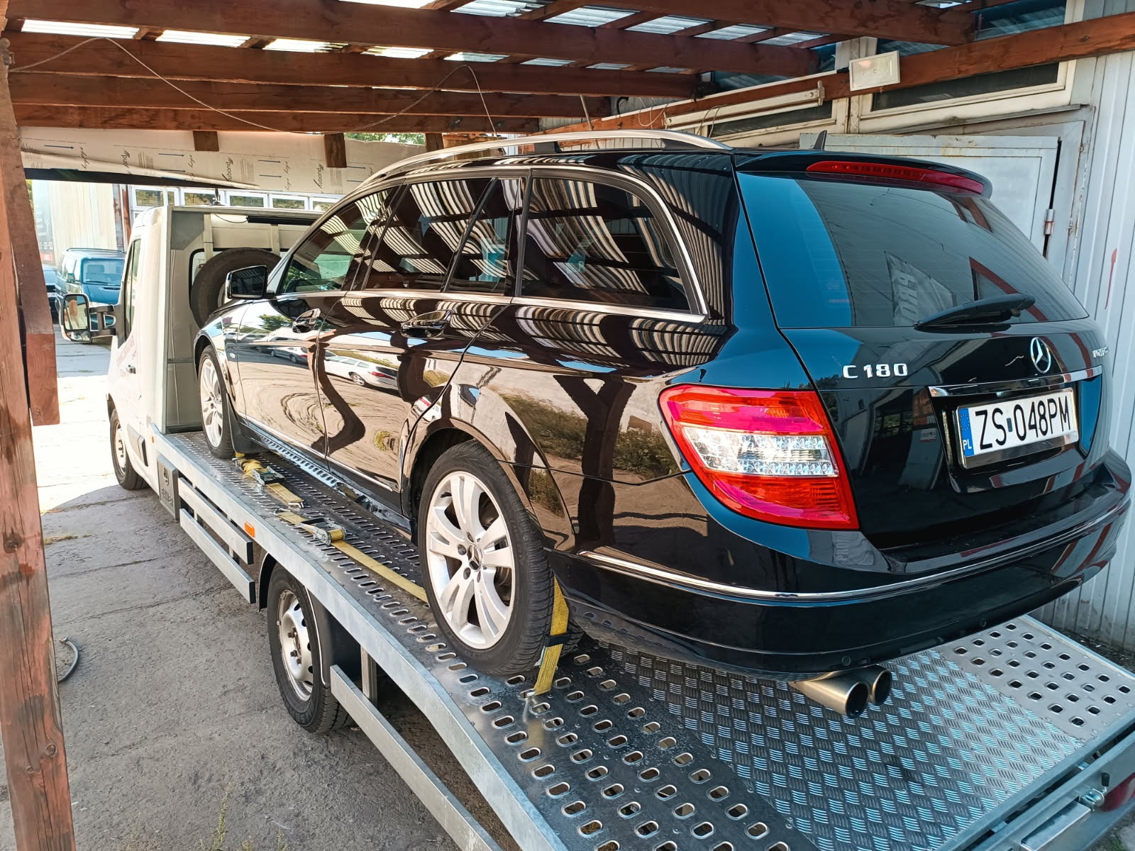 Czarny Mercedes C180 kombi, zabezpieczony pasami, transportowany na lawecie o ażurowej platformie, pod zadaszeniem. Widoczne tablice rejestracyjne.