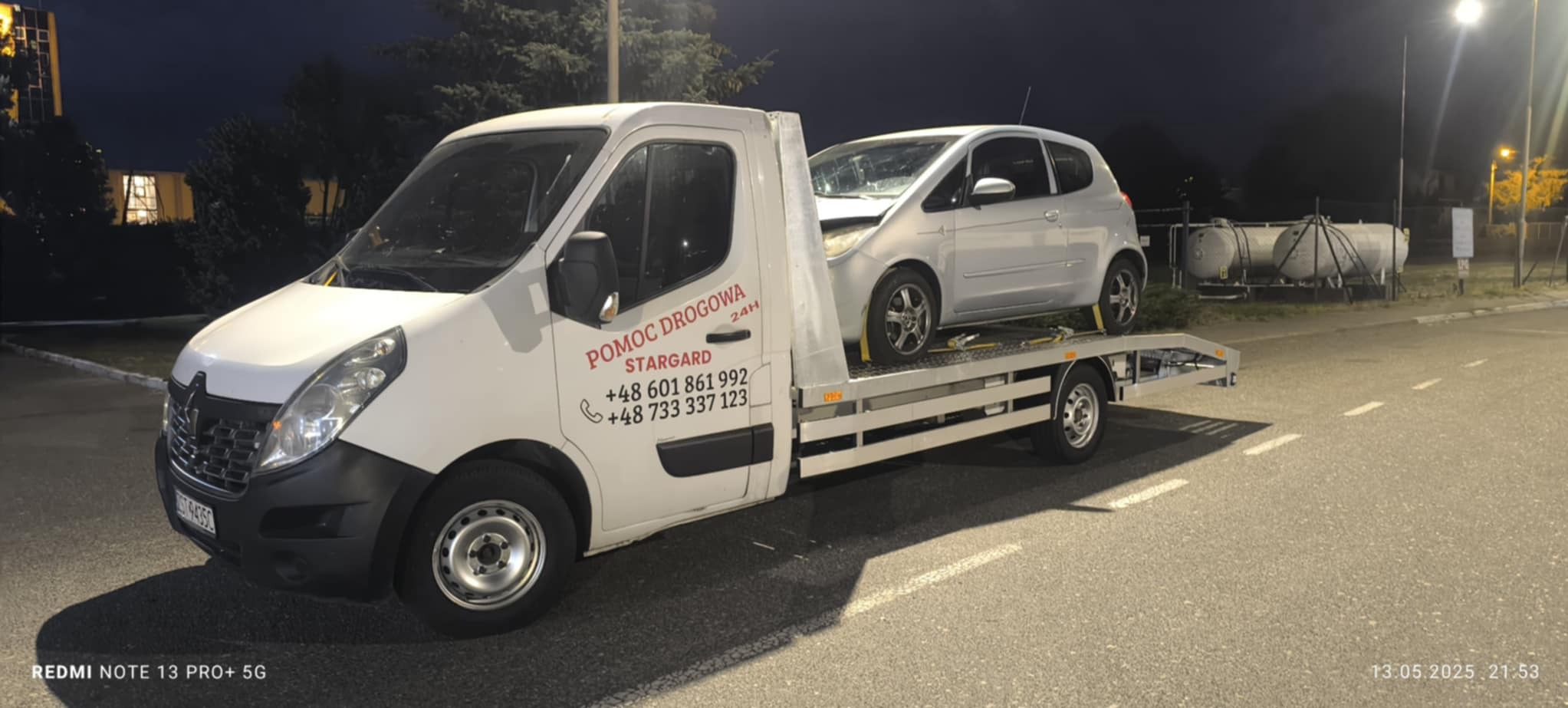 Laweta Stargard z uszkodzonym srebrnym autem na platformie, nocne ujęcie. Widoczny numer telefonu i logo pomocy drogowej na białym samochodzie.