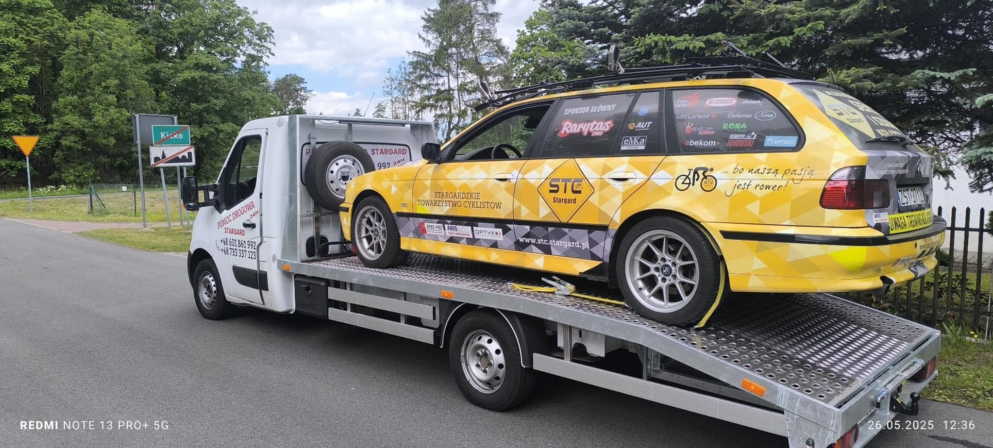 Żółte kombi z rowerami na dachu, oklejone logotypami, transportowane na lawecie pomocy drogowej. Widoczny znak miejscowości Kicko.