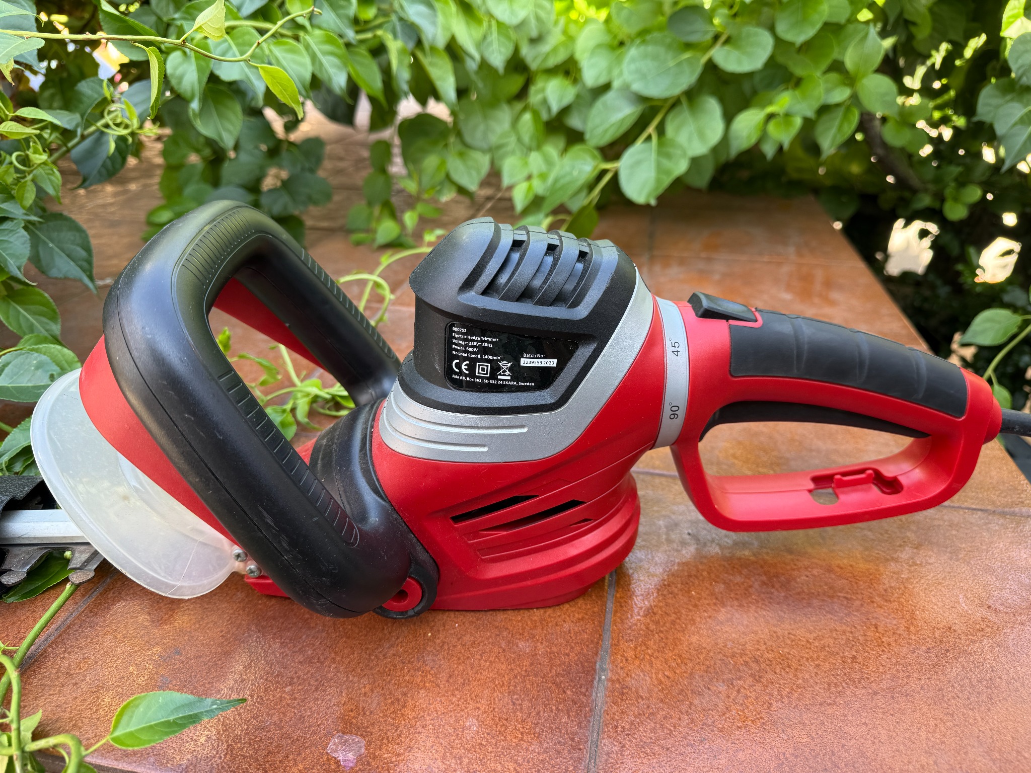 Elektryczne nożyce do żywopłotu w czerwono-czarnej obudowie, leżące na brązowych płytkach, z zielonym żywopłotem w tle. Widoczna osłona ostrzy.
