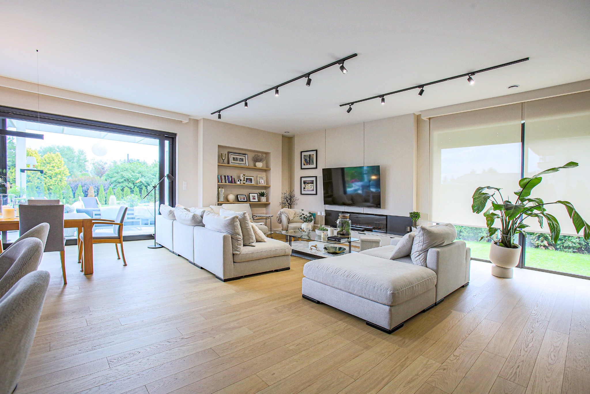 Jasny salon z parkietem, narożnikami i dużym oknem z widokiem na ogród. Minimalistyczny design z oświetleniem szynowym i roślinami doniczkowymi.