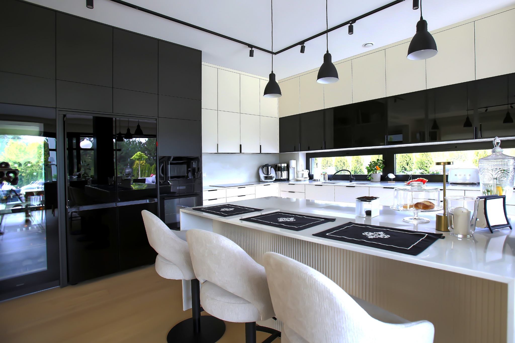 Nowoczesna kuchnia z wyspą: białe blaty, czarne akcenty, krzesła barowe z jasną tapicerką. Widok na ogród przez okno. Minimalistyczny design, eleganckie detale.
