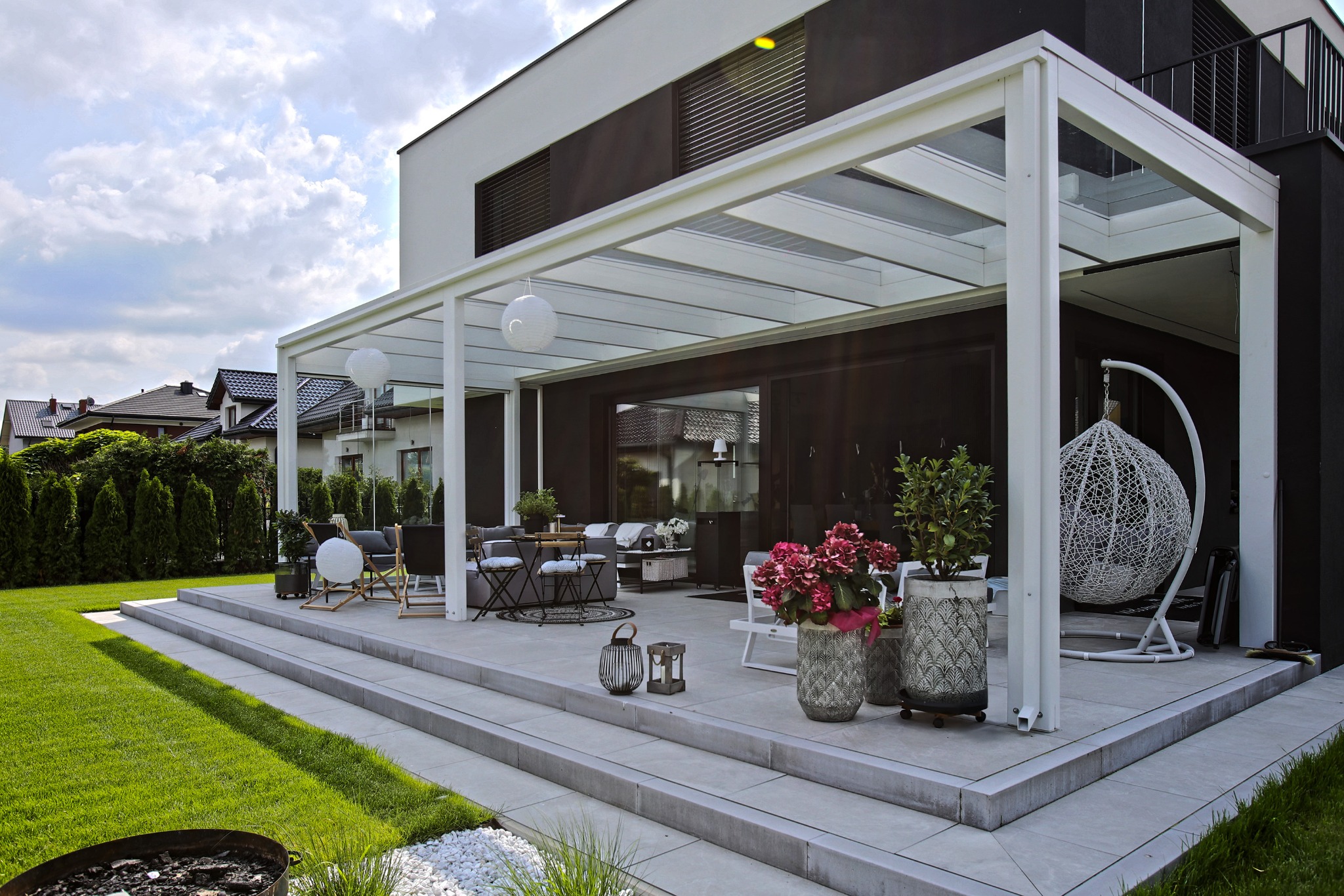 Nowoczesny dom z tarasem z pergolą, wiszącym fotelem i kwiatami w donicach. Architektura minimalistyczna, zadbany trawnik i elementy dekoracyjne tworzą przytulną przestrzeń.