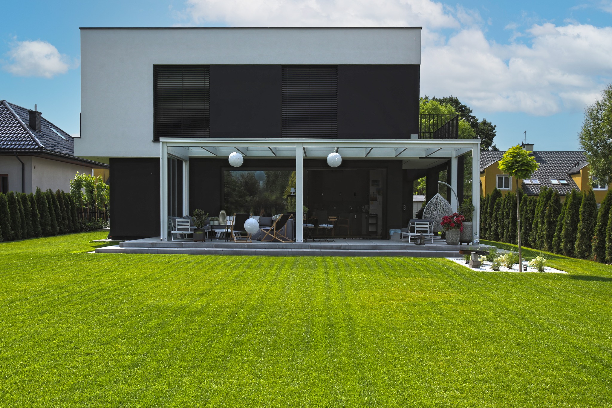 Nowoczesny dom z przeszklonym tarasem i zadbanym trawnikiem. Minimalistyczna architektura z czarno-białą elewacją. Widok na ogród i sąsiednie budynki.