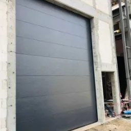 Zamknięta, segmentowa brama garażowa w kolorze antracytu, zamontowana w surowym otworze budynku w trakcie budowy. Widoczne bloczki i stalowa konstrukcja.